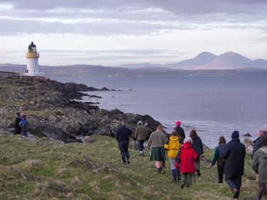 Picture of walkers walking along a shore towards a lighthouse, hills in the distance