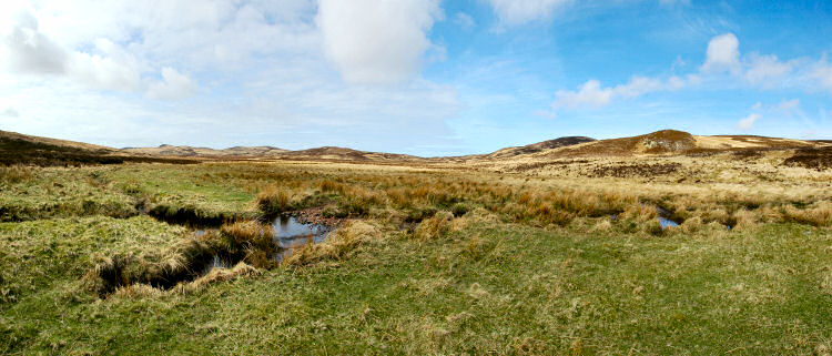Picture of the view over a wide glen with rough ground, a burn winding its way through it