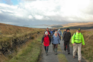 Picture of walkers coming up a single track road, a bay in the background. Dark clouds overhead